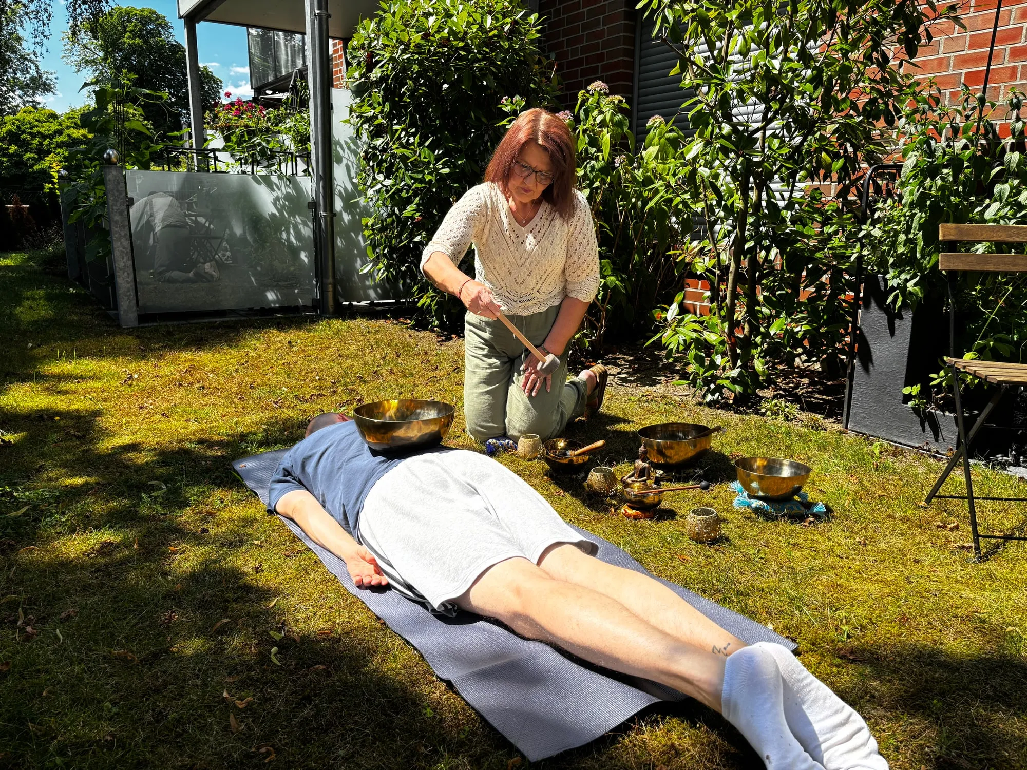 Annette Pitcher kniet neben einer liegenden Person im Garten und schlägt eine Klangschale an